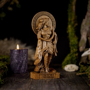 May include: A wooden statue of the Roman goddess Diana, carved in detail. She is depicted standing with a bow and arrow, and her name is inscribed on the base. The statue is set against a dark background with moss and a candle.