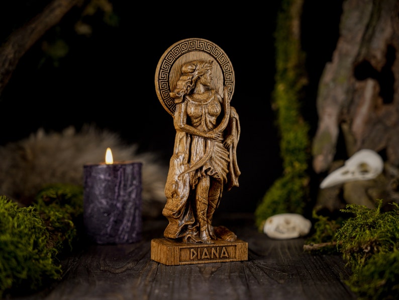 May include: A wooden statue of the Roman goddess Diana, carved in detail. She is depicted standing with a bow and arrow, and her name is inscribed on the base. The statue is set against a dark background with moss and a candle.