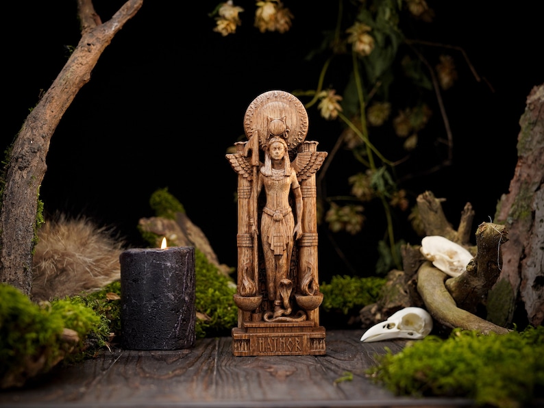 May include: A wooden statue of a goddess with wings, holding a staff and a cup, standing on a pedestal with the word "HATHOR" inscribed on it. The statue is surrounded by moss, branches, and a black candle.