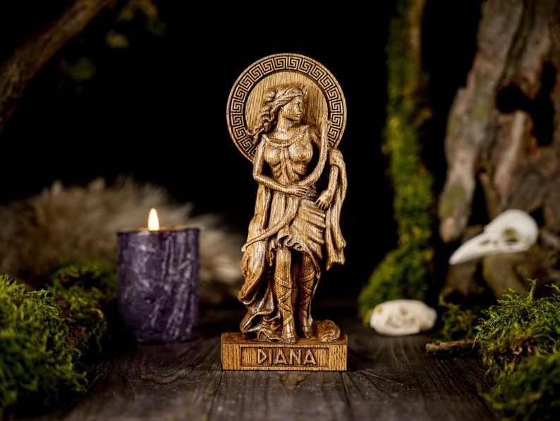 May include: Wooden statue of the Roman goddess Diana, standing with a bow and arrow, with a Greek key design around her head. The statue is on a wooden base with the word "DIANA" inscribed on it.