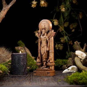 May include: A wooden statue of a goddess with wings and a crown, standing on a pedestal with a base. The statue is surrounded by moss, branches, and a black candle.