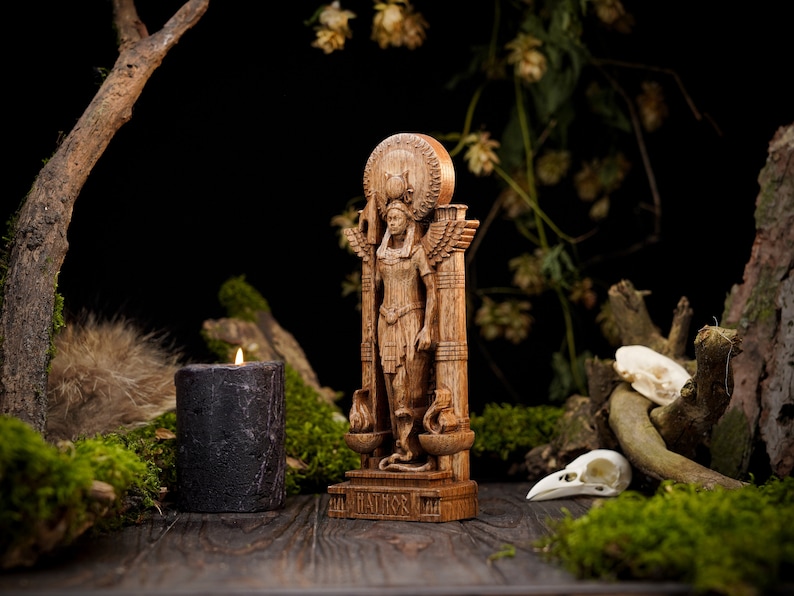 May include: A wooden statue of a goddess with wings and a crown, standing on a pedestal with a base. The statue is surrounded by moss, branches, and a black candle.