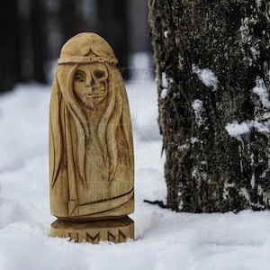 Puede incluir: Una talla de madera de una mujer con una cara de calavera. La talla est&aacute; de pie en la nieve con un tronco de &aacute;rbol en el fondo. La talla tiene la palabra "JMMN" tallada en la base.