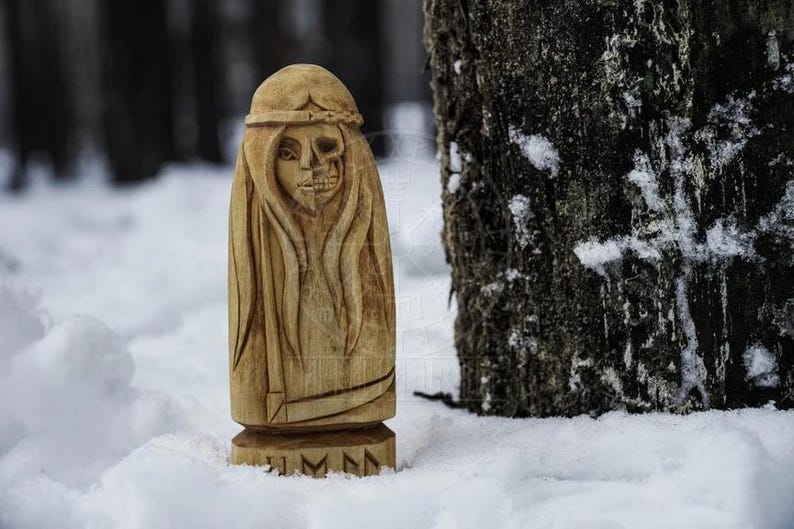 Puede incluir: Una talla de madera de una mujer con una cara de calavera. La talla est&aacute; de pie en la nieve con un tronco de &aacute;rbol en el fondo. La talla tiene la palabra "JMMN" tallada en la base.