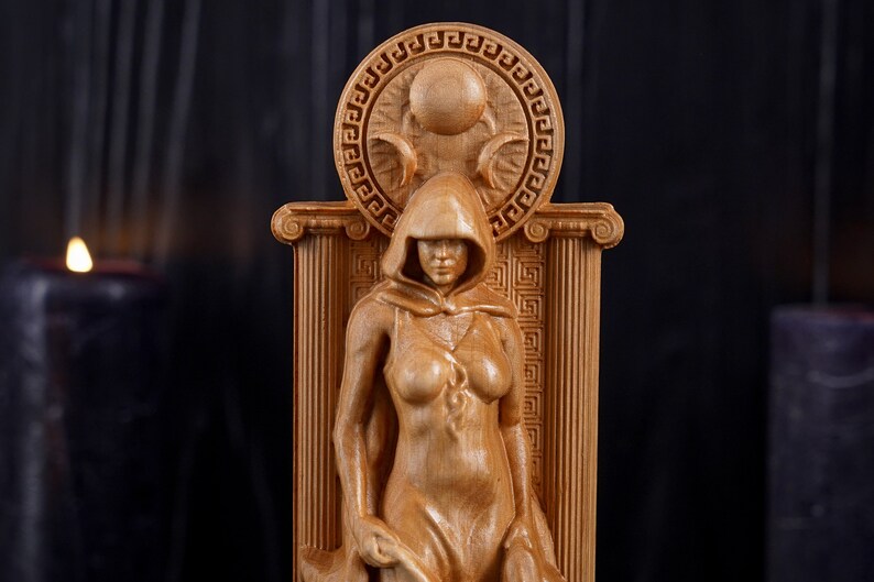 Puede incluir: Una escultura de madera de una mujer con una t&uacute;nica con capucha, de pie en una puerta con un dise&ntilde;o circular sobre su cabeza. La escultura est&aacute; tallada en madera de color marr&oacute;n claro.