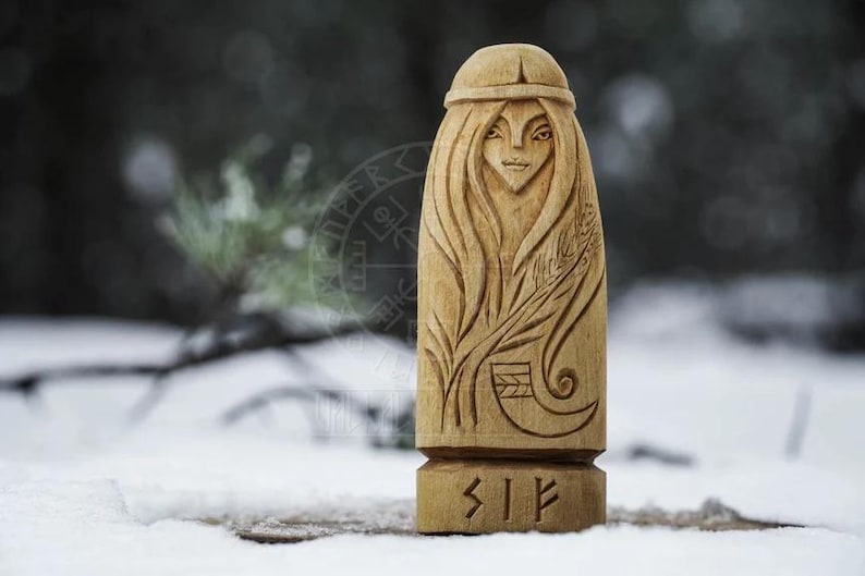 Puede incluir: Una talla de madera de una diosa n&oacute;rdica con el pelo largo y un tocado. La talla est&aacute; detallada con una planta y un s&iacute;mbolo de runa, "Sif", en la base.