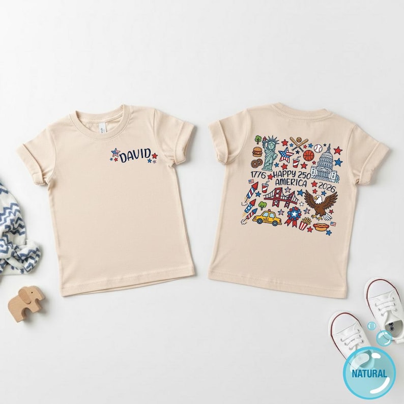 Puede incluir: Dos camisetas beige con dise&ntilde;os patri&oacute;ticos. Una camiseta tiene el nombre "DAVID" con detalles de estrellas. La otra camiseta presenta una ilustraci&oacute;n colorida con el texto "1776 HAPPY 250 AMERICA 2026" y varios s&iacute;mbolos americanos.