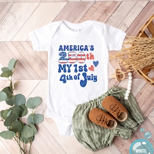以下が含まれることがあります： 「America's 250th My 1st 4th of July」と赤と青で書かれた白いベビーボディスーツ。緑のストライプのショートパンツと茶色のベビーシューズがセットになっています。おしゃぶりと装飾的な要素も含まれています。