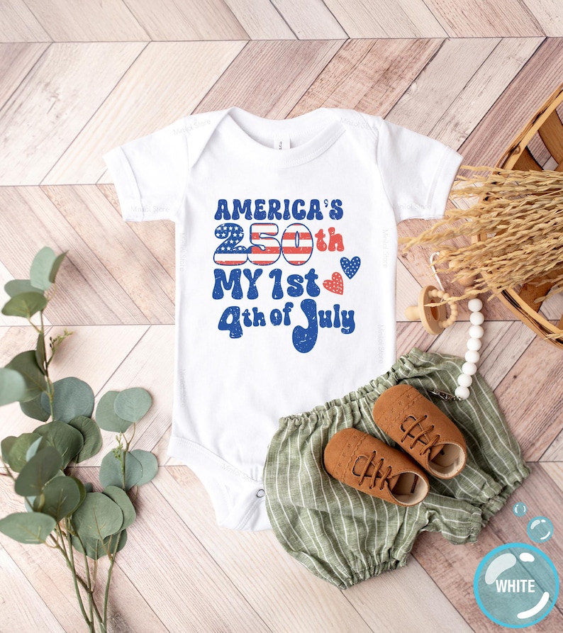 以下が含まれることがあります： 「America's 250th My 1st 4th of July」と赤と青で書かれた白いベビーボディスーツ。緑のストライプのショートパンツと茶色のベビーシューズがセットになっています。おしゃぶりと装飾的な要素も含まれています。