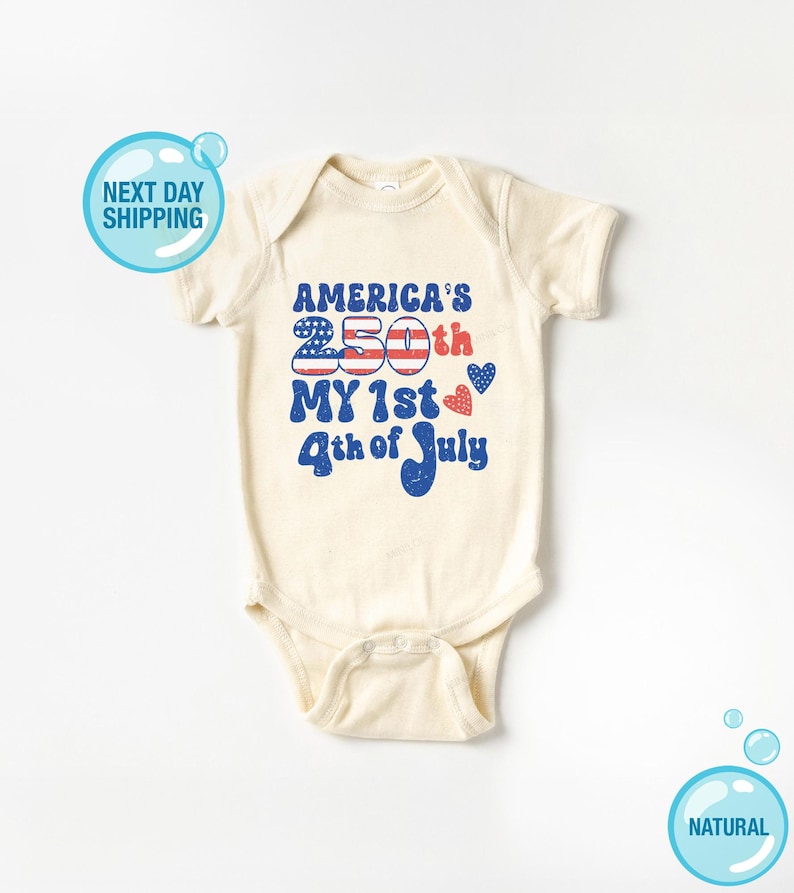 以下が含まれることがあります： クリーム色のベビーボディスーツ。赤、白、青で「America's 250th My 1st 4th of July」の文字がプリントされています。デザインには、様式化されたアメリカ国旗とハートの形が含まれています。柔らかい素材で作られています。