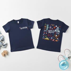 Puede incluir: Camisetas azul marino de manga corta. Una camiseta tiene el nombre "HUDSON" en una fuente colorida. La otra camiseta presenta un gr&aacute;fico con el texto "1776 HAPPY 250 AMERICA 2026" y s&iacute;mbolos patri&oacute;ticos.