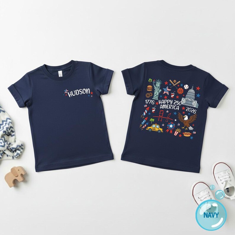 Puede incluir: Camisetas azul marino de manga corta. Una camiseta tiene el nombre "HUDSON" en una fuente colorida. La otra camiseta presenta un gr&aacute;fico con el texto "1776 HAPPY 250 AMERICA 2026" y s&iacute;mbolos patri&oacute;ticos.