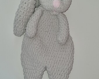 Könnte beinhalten: Ein grauer Häkelhase mit blauen Augen und einer rosa Nase. Der Hase liegt auf der Seite mit ausgestreckten Beinen.