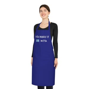 Op de afbeelding: Een koningsblauwe schort met de tekst "FETA MAKES IT BETTER" en een Griekse vlag. De schort wordt gedragen door een persoon in een zwart shirt met lange mouwen.