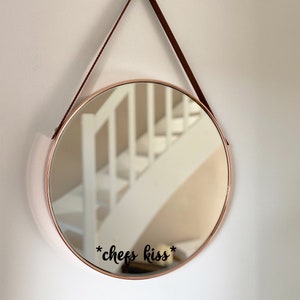 May include: Circular mirror with a copper-toned frame, suspended by a brown leather strap. The mirror reflects a white staircase. The words "*chefs kiss*" are written in black script on the mirror's surface.