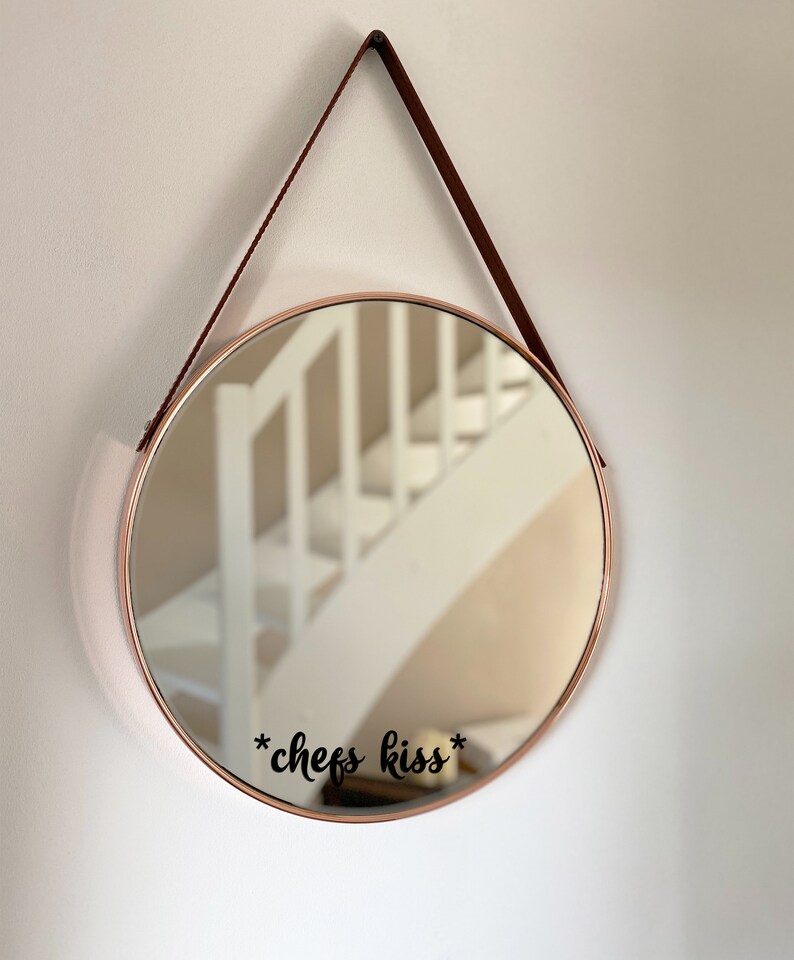 May include: Circular mirror with a copper-toned frame, suspended by a brown leather strap. The mirror reflects a white staircase. The words "*chefs kiss*" are written in black script on the mirror's surface.