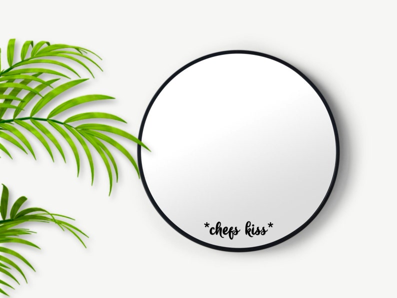 May include: A circular mirror with a black frame, featuring the phrase "*chefs kiss*" in black script. The mirror is set against a white backdrop, with green palm leaves in the upper left corner.