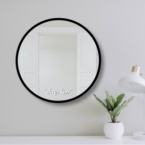 May include: A circular mirror with a black frame, featuring the text "*chef's kiss*" in white script. The mirror reflects a white doorway. A white desk lamp and a small potted plant sit on a white surface.