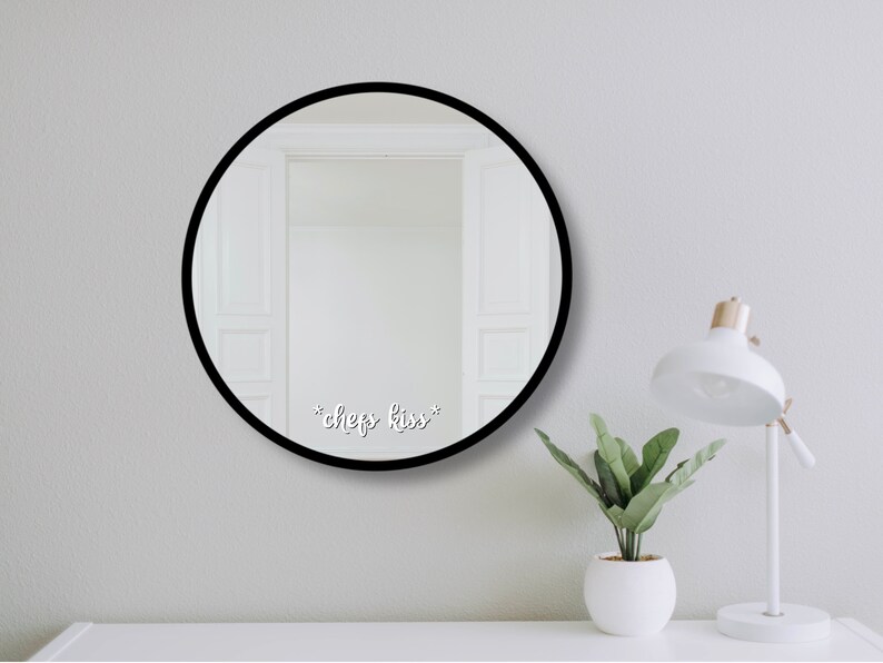 May include: A circular mirror with a black frame, featuring the text "*chef's kiss*" in white script. The mirror reflects a white doorway. A white desk lamp and a small potted plant sit on a white surface.