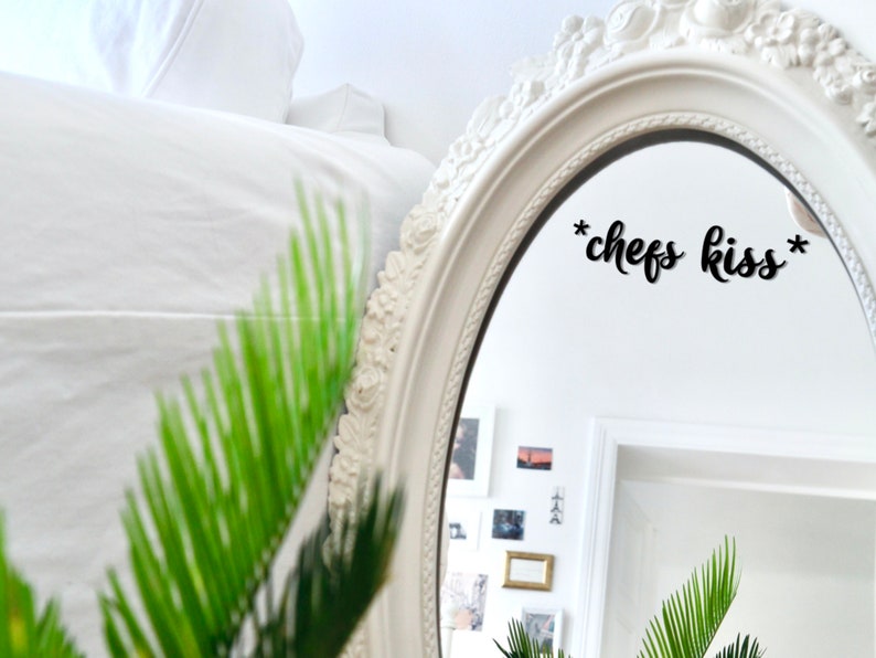 May include: An oval white mirror with an ornate frame, reflecting a bright room. The mirror has the phrase "*chefs kiss*" in black script. Green foliage is visible in the foreground.