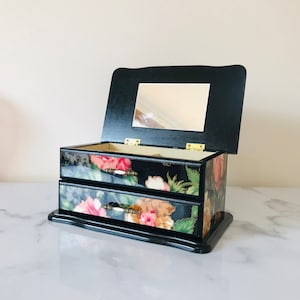 May include: Black wooden jewellery box with a floral print and two drawers. The box has a mirror inside the lid.