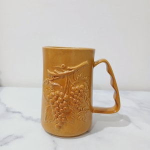 May include: A tan ceramic mug with a large handle. The mug has a raised relief design of grape vines and bunches of grapes. The mug is a medium size and has a smooth, glossy finish.