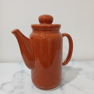 May include: A glazed, terracotta-coloured ceramic teapot with a lid and a curved handle. The teapot features a long spout and a rounded body. The design is simple and classic, suitable for serving tea or other hot drinks.