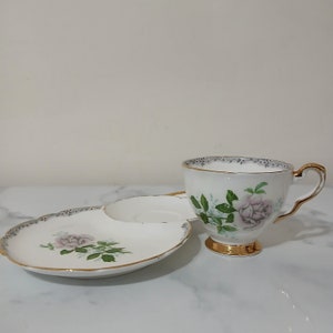May include: A white teacup and saucer with a floral design. The cup and saucer have a gold trim and feature a pink rose with green leaves.