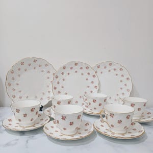 May include: A classic tea set comprising white porcelain cups, saucers, and plates. The set is decorated with a floral design in pink hues and gold edging. The plates have a decorative, scalloped edge.