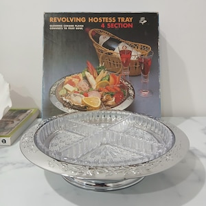 May include: A silver revolving hostess tray with four sections. The tray features a decorative floral design around the edge and a clear plastic insert with four compartments. The tray is shown on a white surface with a box in the background. The box has a picture of the tray with food on it and the text "Revolving Hostess Tray 4 Section Gleaming Chrome Plated Converts to Fruit Bowl".