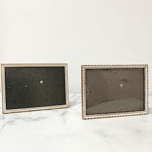 May include: Two rectangular picture frames with a light cream-coloured finish and a delicate beaded texture along the inner and outer edges. Each frame features a dark, solid-coloured backing material and a clear glass front panel for display.