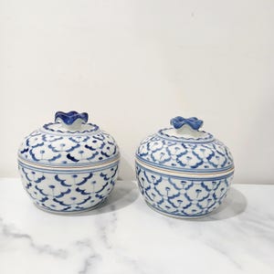 May include: Two matching white and blue ceramic bowls with lids. The bowls feature a floral pattern and a blue, flower-shaped handle on the lid. The bowls are round and sit on a white surface.