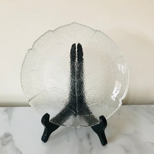 May include: A clear glass leaf-shaped plate with textured detailing, displayed on a black stand. The plate's design mimics the veins of a leaf. The stand is made of black wood.