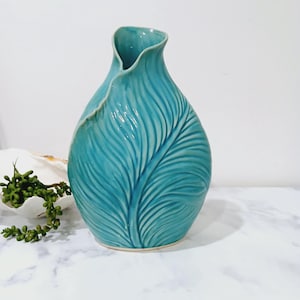 May include: A turquoise ceramic vase with a leaf design. The vase has a unique shape with a wide base and a narrow, curved opening. The leaf pattern is detailed and textured. The vase is a decorative item, suitable for displaying flowers or as a standalone piece.