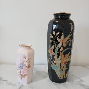 May include: Two decorative vases. A small, pale pink vase with floral designs in blue, pink, and brown. A larger, black vase with gold trim and a floral pattern in orange, brown, and teal.