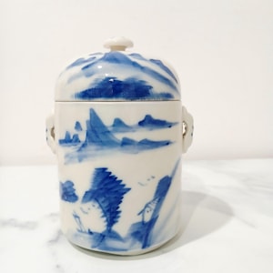 May include: A white ceramic jar with a lid, decorated with a blue and white landscape design. The jar has a rounded shape, two small handles, and a small knob on the lid. The landscape design includes mountains and trees.