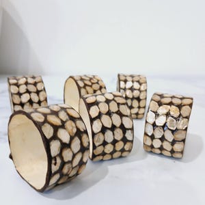 May include: A set of six decorative napkin rings. Each ring is cylindrical, with a dark brown exterior and a light beige interior. The exterior is covered in small, irregularly shaped, light beige pieces.