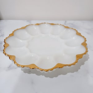 May include: White, scalloped-edge serving dish with a gold rim. The dish has a central area with several rounded indentations, likely for holding eggs. The gold rim adds a touch of elegance to the serving piece.