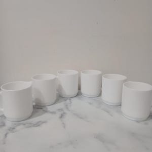 May include: Six white ceramic mugs with a simple, cylindrical design. Each mug has a handle and a slightly flared base. The mugs are arranged on a marble-patterned surface against a plain white background. The mugs are all the same size and shape.