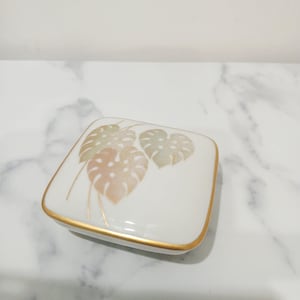 May include: A square white porcelain trinket box with rounded corners and a gold-trimmed edge. The lid features a delicate, soft-toned illustration of three monstera leaves in muted shades of peach, sage green, and light brown with gold accents.