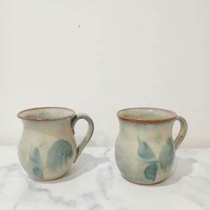 May include: Two ceramic mugs with a rounded, bulbous shape and a handle on the side. The mugs feature a cream-coloured base glaze with soft, blended blue-green leaf-like patterns and a distinct brown rim around the top edge of each vessel.