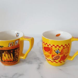 May include: Two yellow ceramic mugs with white interiors. One mug features a fireplace design, the other has a checkered pattern with pumpkins and a butterfly. Both mugs have a handle and the Lipton logo.