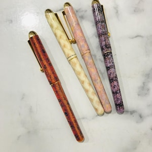 May include: Four fountain pens with different patterned barrels. The pens are gold-toned and have a classic design. The patterns include a red and orange marbled design, a pink and white marbled design, a light pink and white marbled design, and a purple and black marbled design.