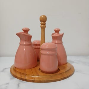 May include: A pink ceramic condiment set on a wooden base, featuring two oil dispensers, two shakers, and a salt shaker, with a central wooden handle. The round wooden base has a raised edge. The set is designed for the table.