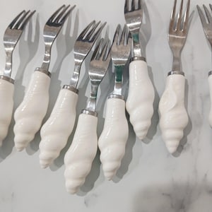 May include: A set of dessert forks featuring stainless steel tines and white, seashell-shaped handles. The handles have a glossy finish and are attached to the tines with a silver metal band. The forks are arranged in a slightly fanned-out pattern.