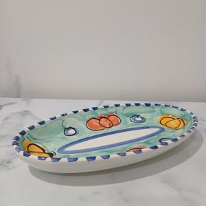 May include: An oval ceramic serving dish with a blue and white striped border. The dish is decorated with hand-painted green, orange, and yellow abstract shapes.