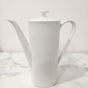 May include: A tall white porcelain coffee pot featuring a cylindrical body with vertical ribbed detailing, a long slender spout, a curved handle, and a matching lid with a small knob handle on top, presented in a clean and simple design.