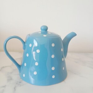 May include: A light blue ceramic teapot featuring a repeating pattern of white polka dots. The teapot has a curved handle, a matching lid with a small knob handle, and a short spout. The surface is smooth and glossy with a rounded body shape.