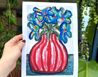 May include: A colorful painting of blue flowers in a red vase. The vase has vertical white stripes. The artwork is on white paper and held by a hand. The background includes plants and a striped lampshade.