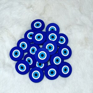 May include: A collection of blue and white evil eye charms. Each charm features a blue background with a white and black eye design. The charms are round and are arranged on a white, fluffy surface.